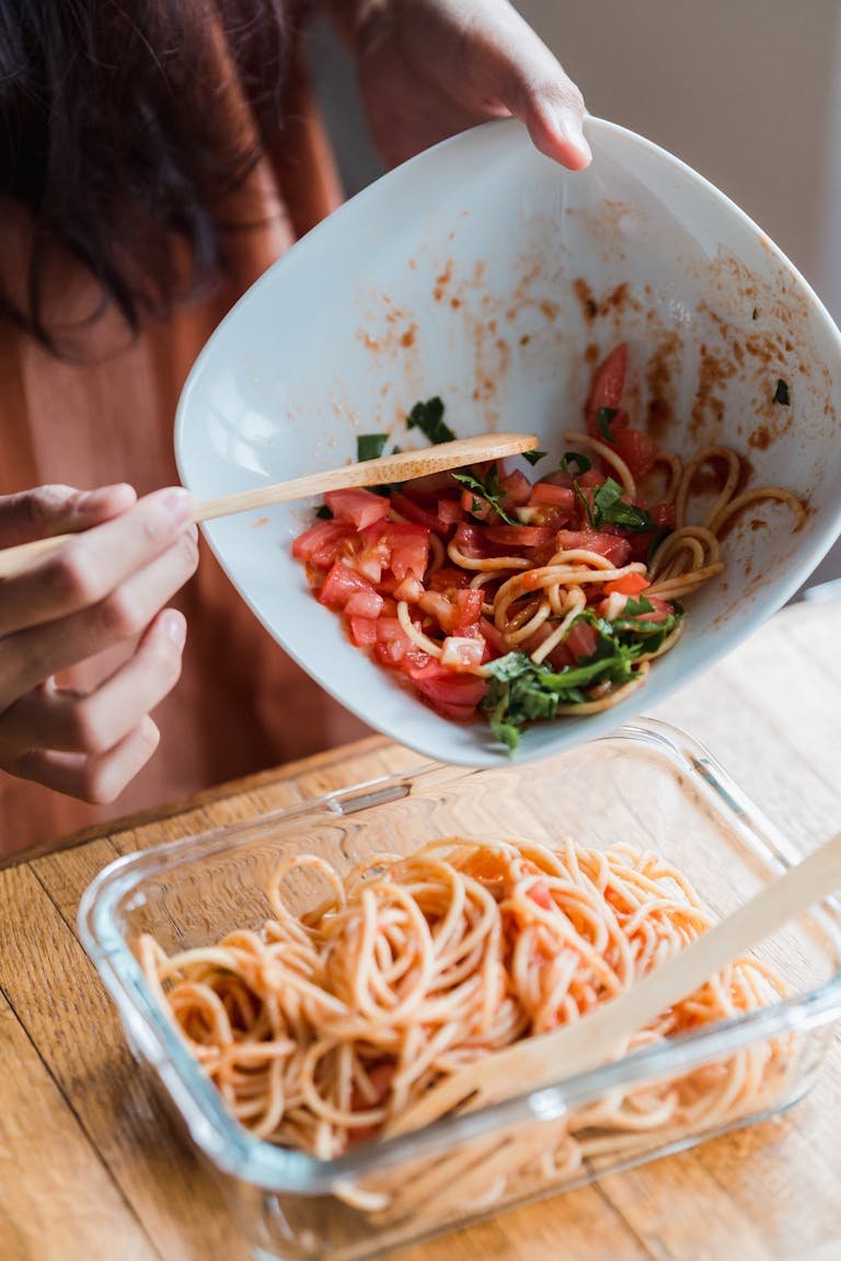 A person adds tomatoes to pasta in a glass dish on a wooden table.