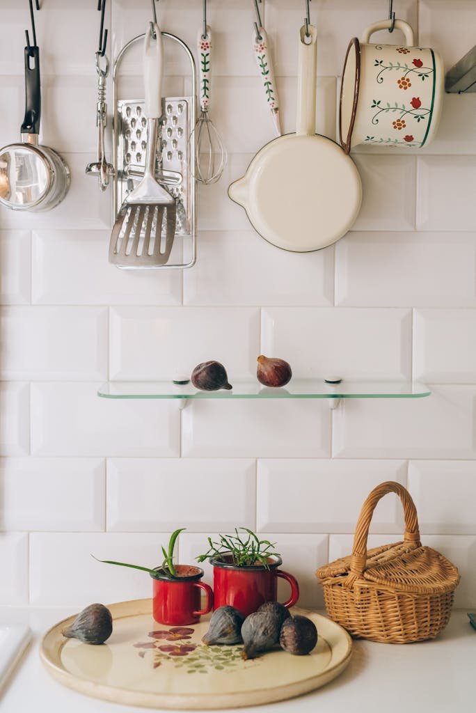 Charming kitchen scene with wall-mounted pot racks, hanging utensils, red mugs, figs, and a wicker basket on white tile wall.