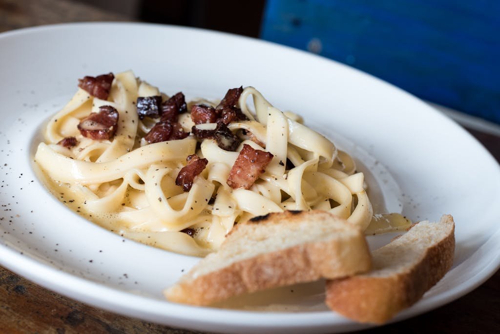 Close-up of creamy pasta carbonara topped with bacon, served with fresh bread.