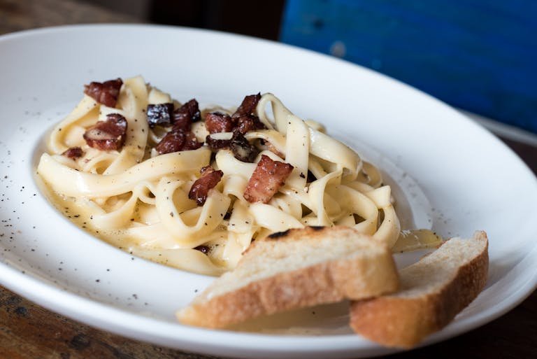Close-up of creamy carbonara pasta topped with bacon, served with fresh bread in a restaurant.