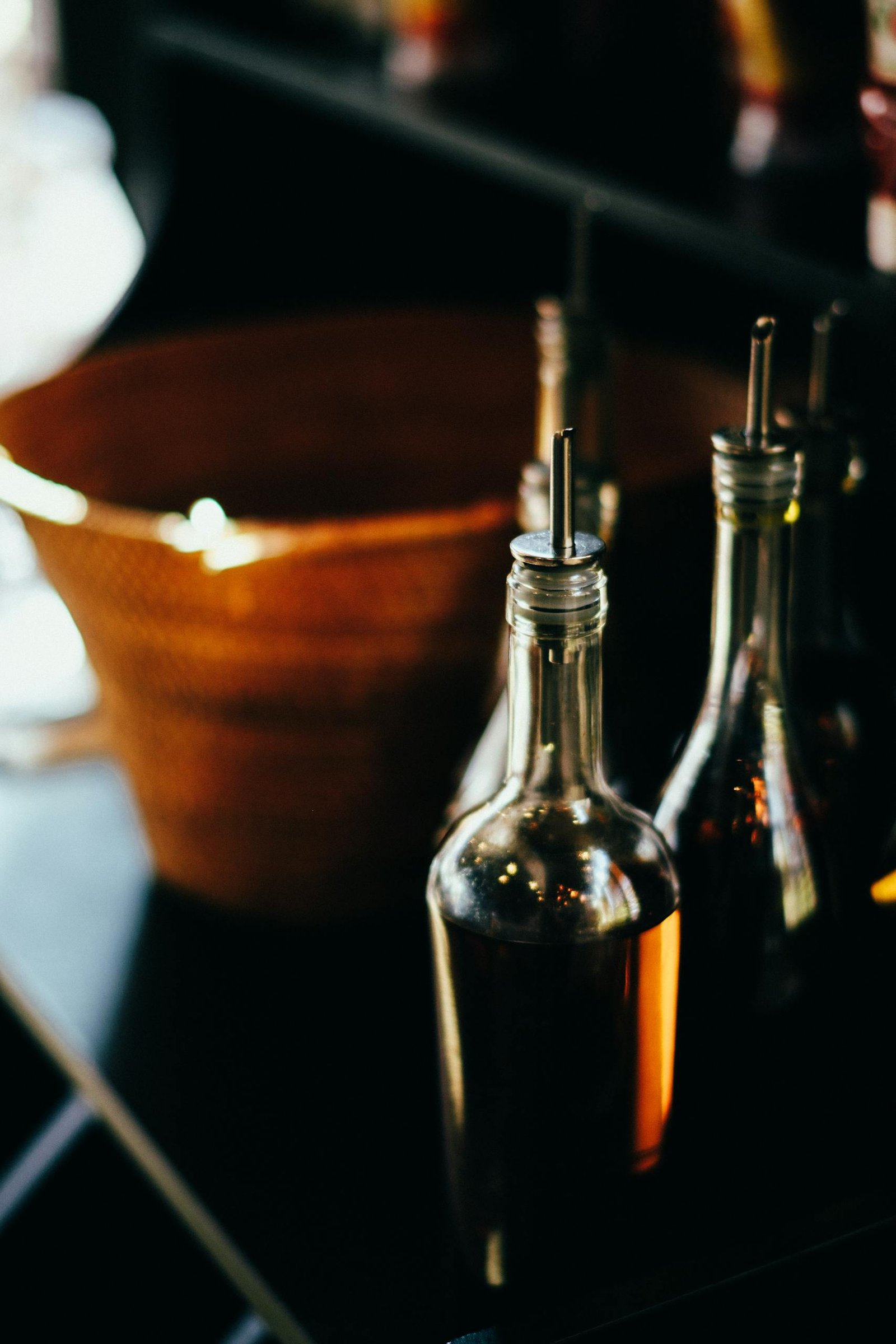Close-up shot of clear glass bottles filled with balsamic vinegar and olive oil, on a dark counter beside a wicker basket.