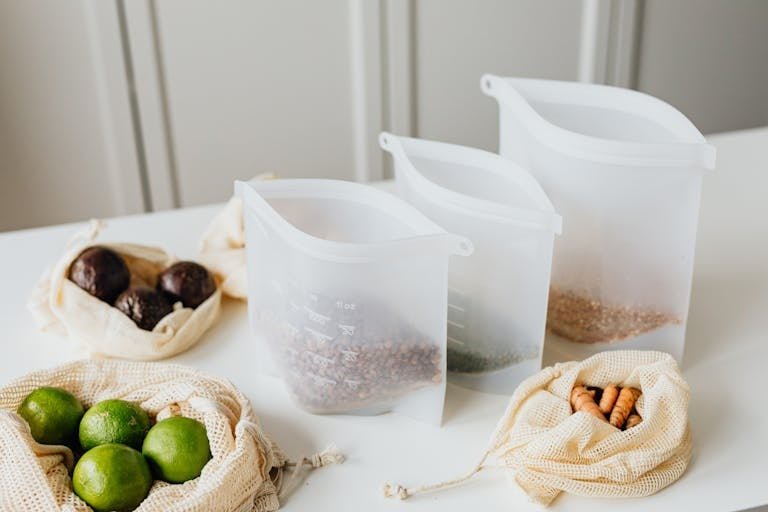 Divided silicone food storage bag showing reusable silicone and cloth bags storing lime, turmeric, and grains on a kitchen table.