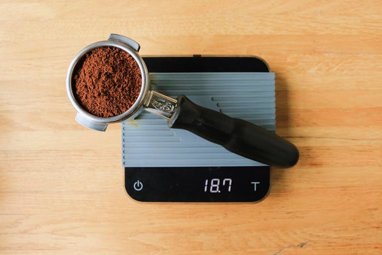 Top view of kitchen gadgets. A portafilter filled with ground coffee resting on a digital scale on a wooden table.