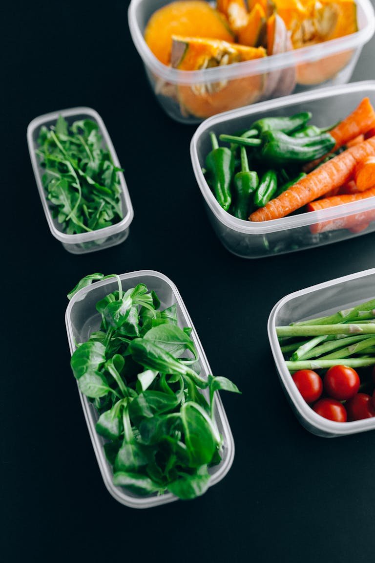 Assorted fresh vegetables including arugula, carrots, and tomatoes in eco-friendly healthy meal prep containers against a black background.