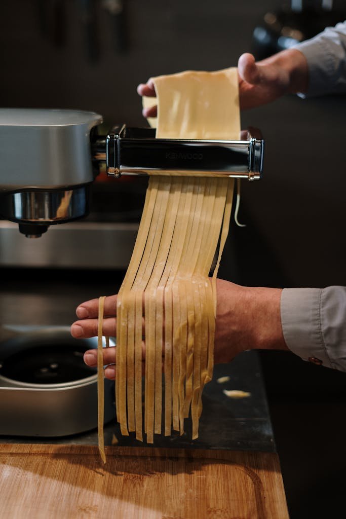 Hands making fresh pasta with an electric pasta machine in a kitchen.