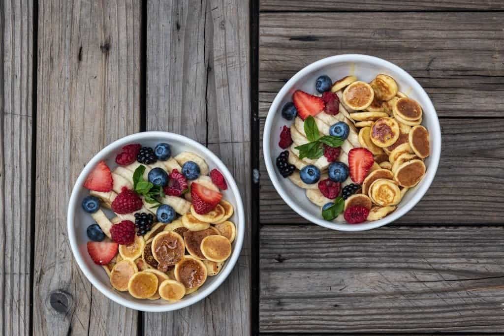 Top view of two bowls with mini pancakes and fresh fruits on rustic wood table.