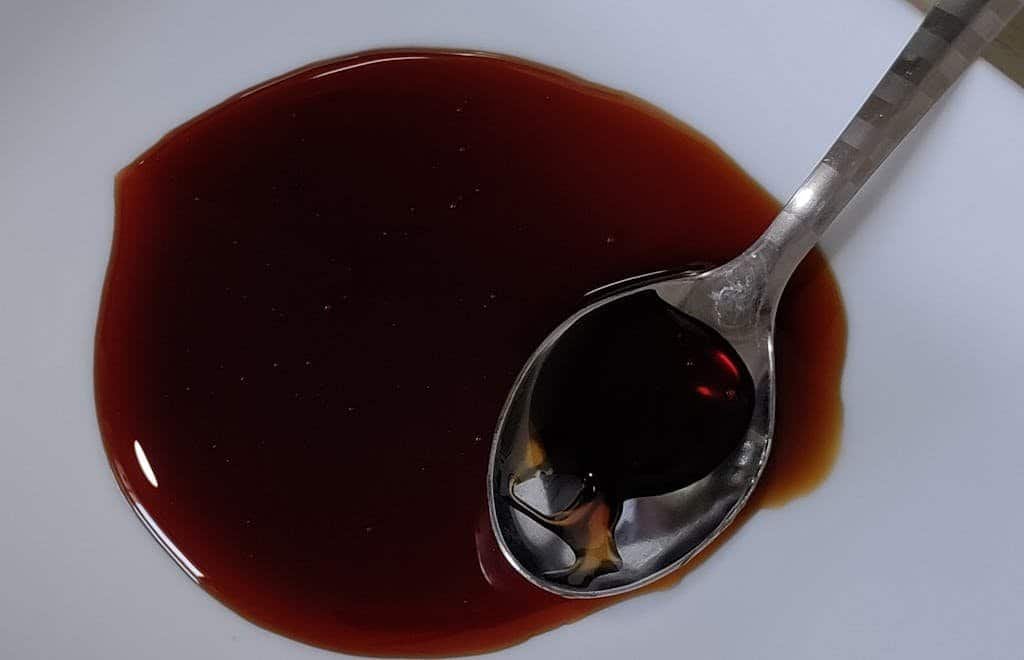 A close-up image of a spoon holding dark soy sauce on a white ceramic plate.