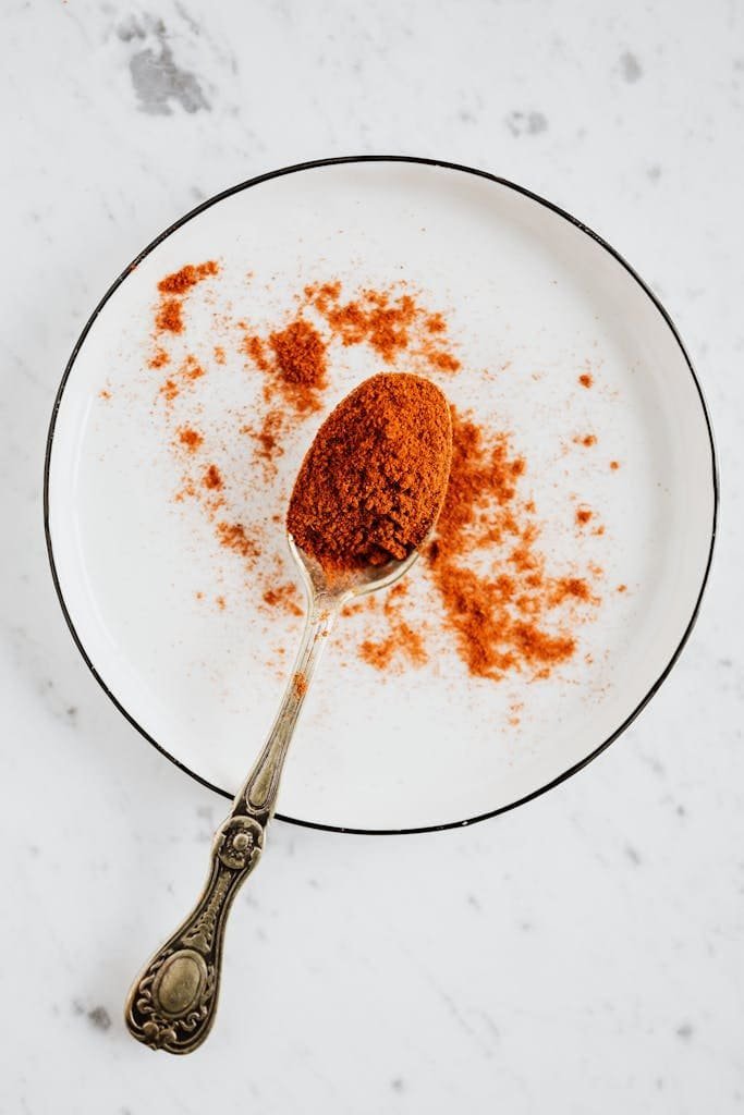 Top view of metal spoon with ground paprika in metal round bowl on marble desktop