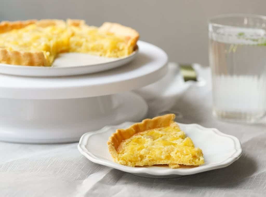 A delicious lemon tart with a slice served on a white plate beside a glass of water.