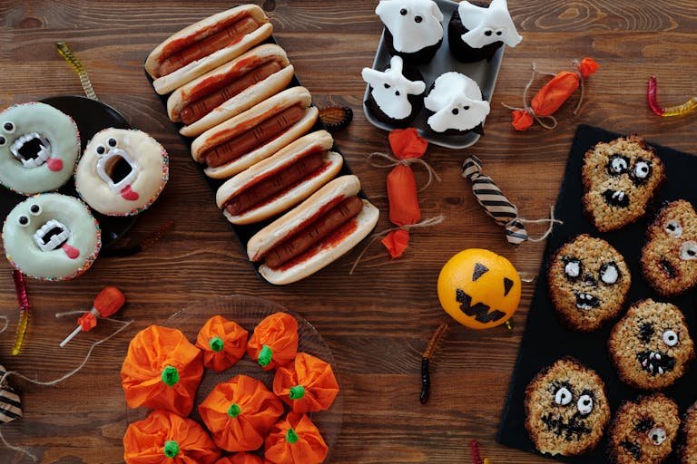 Festive Halloween treats including ghost cupcakes, spooky hot dogs, and monster donuts on a wooden table.
